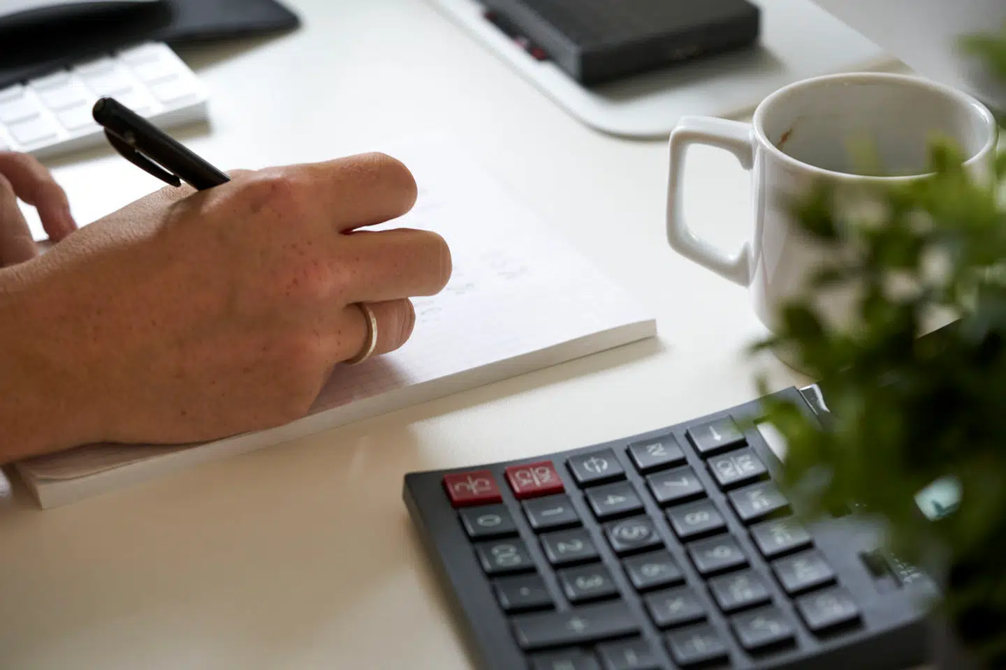 Hånd skriver på en blok på et bord med lommeregner, kaffekop og plante, København den 22. juli 2021