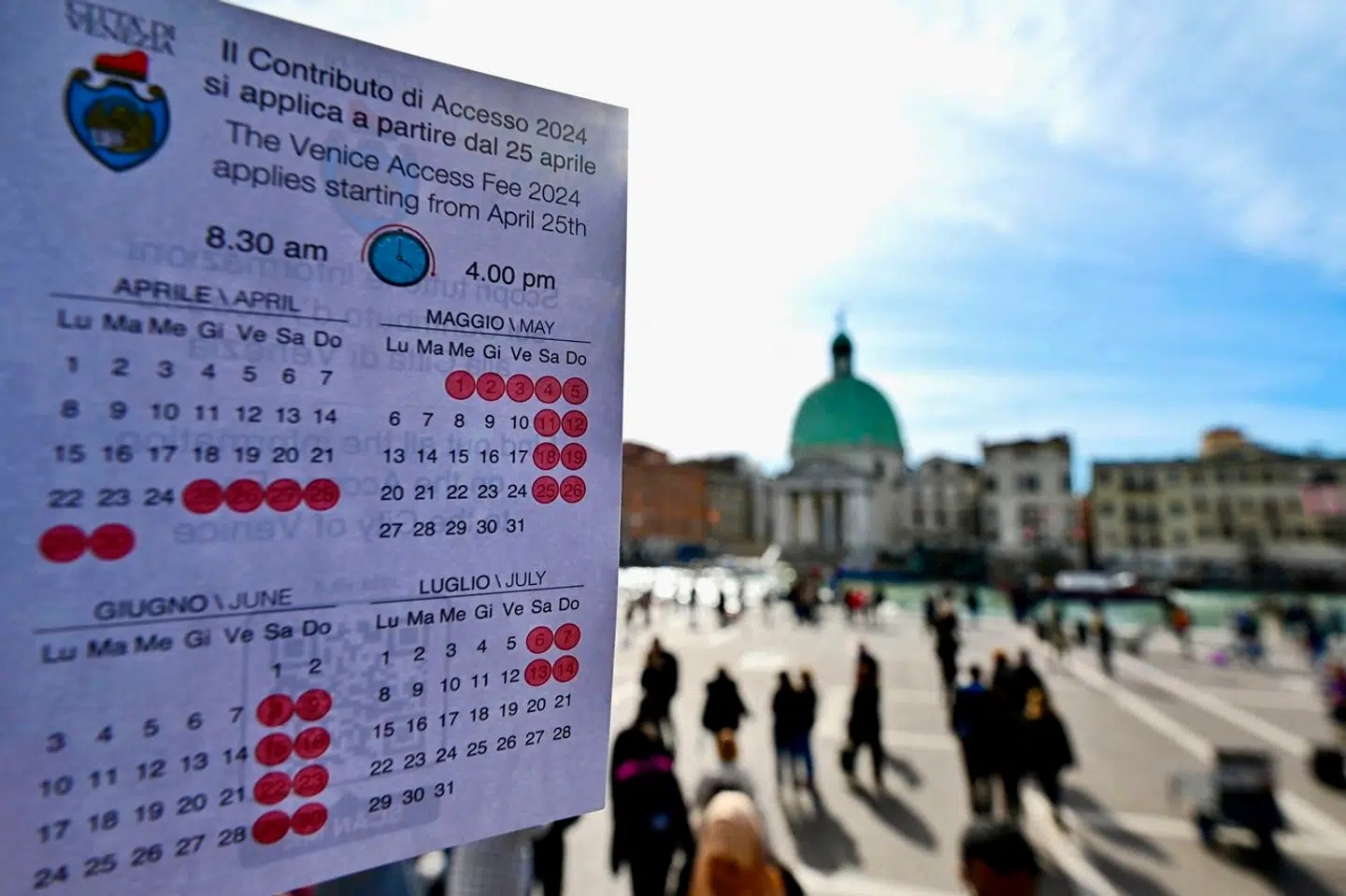 En person viser en kalender, hvor det fremgår, hvilke dage der skal betales entré for at besøge Venedig som endagsturist. I baggrunden ses Santa Lucia-togstationen.
