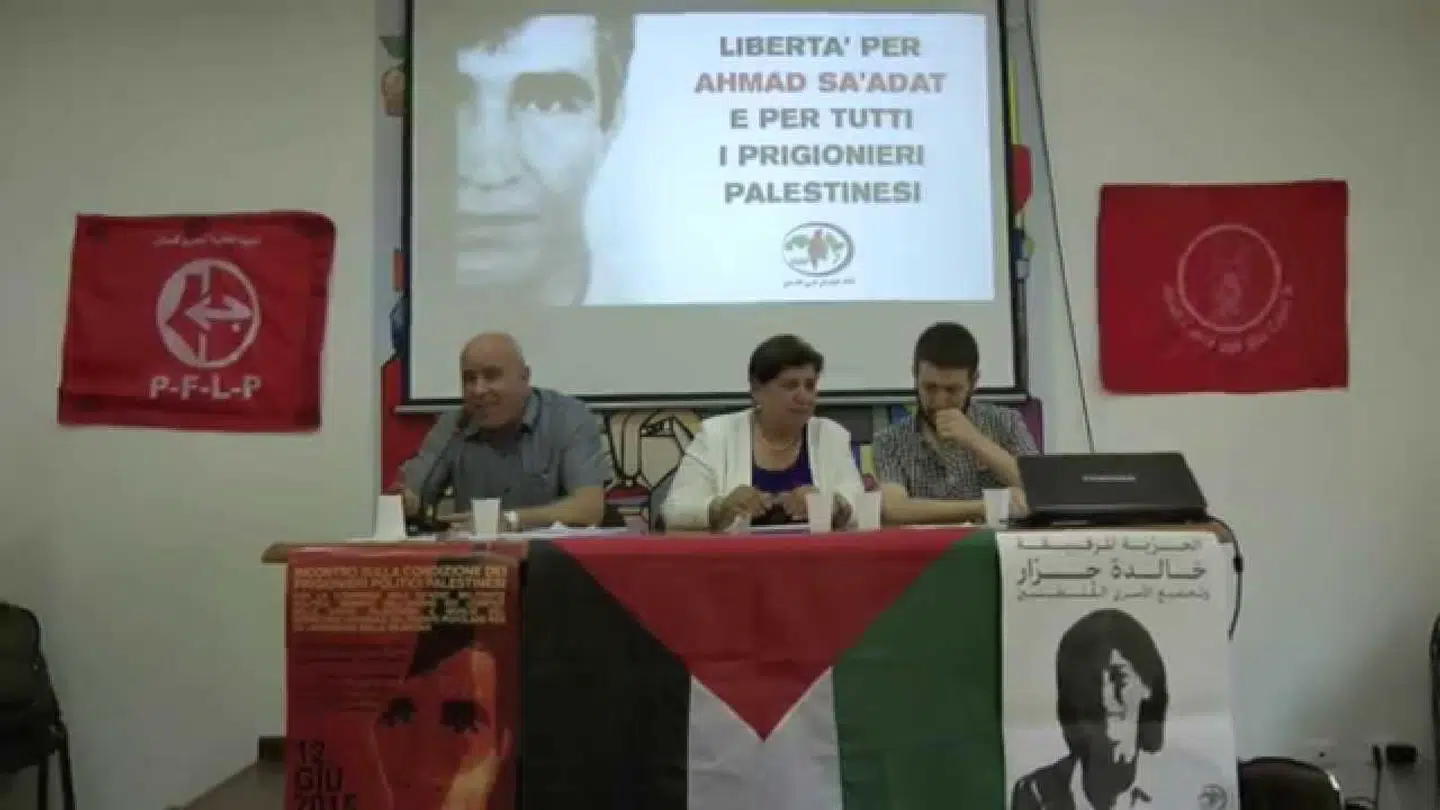 Her ses Abla Sa'adat holde foredrag i Italien. Bag hende er et billede af hendes mand, Ahmad Sa'adat, samt et PFLP-banner.