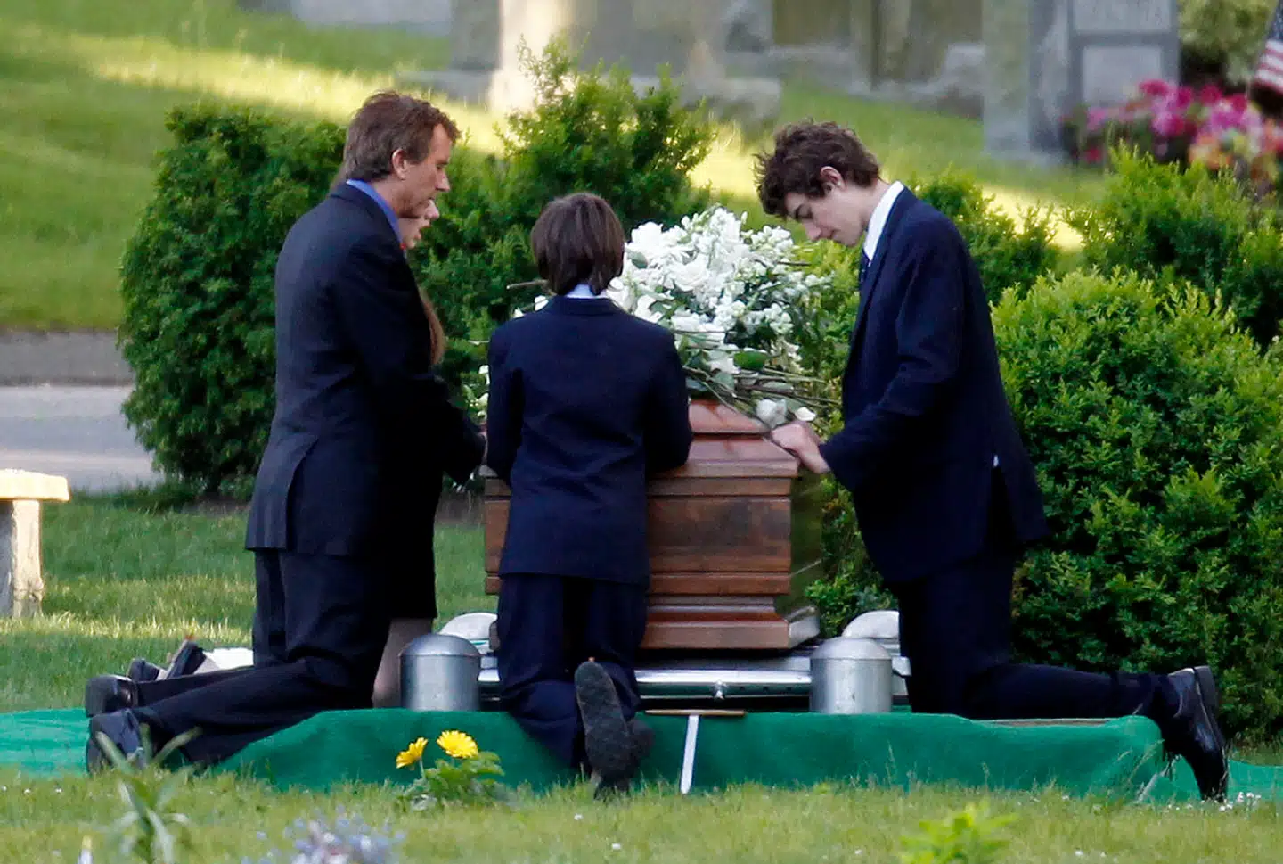 Virkeligheden, som Kennedy-familien gerne ville have den til at se ud – Robert F. Kennedy Jr. (tv.) knæler ved siden af kisten med sin afdøde kone, som han støttede gennem hendes ulykke. Ved siden af er parrets børn. Virkeligheden var en helt anden, og selv gravstedet var fup.