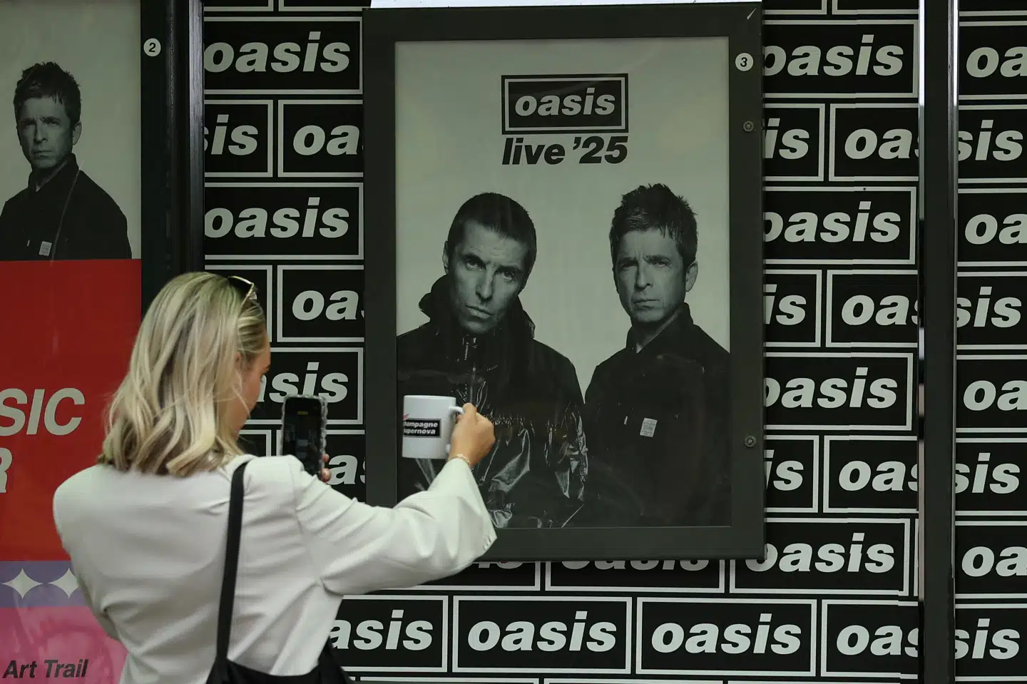 En kvinde tager et billede ved en plakat med brødrene Gallagher, der er frontfigurer i det gendannede Oasis. Bandet er på turné og spiller på udsolgte stadioner. Lørdag aften var der koncert på Wembley i London, hvor en mand døde efter et fald. (Arkivfoto)