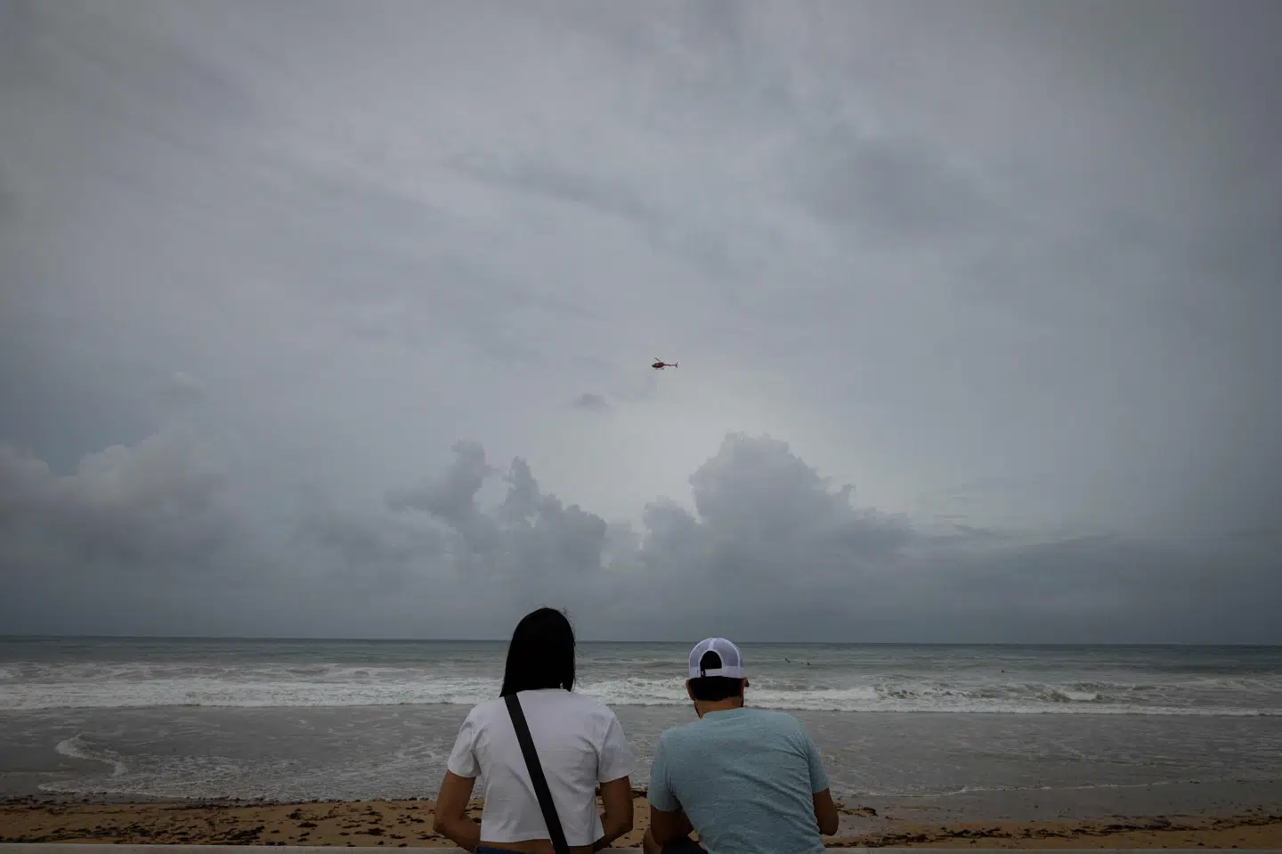 En helikopter flyver over en strand, mens et par ser på i Luqillo i Puerto Rico lørdag. Orkanen Erin ventes at medføre nedbør i Caribien, men ventes ikke at nå fastlandet.