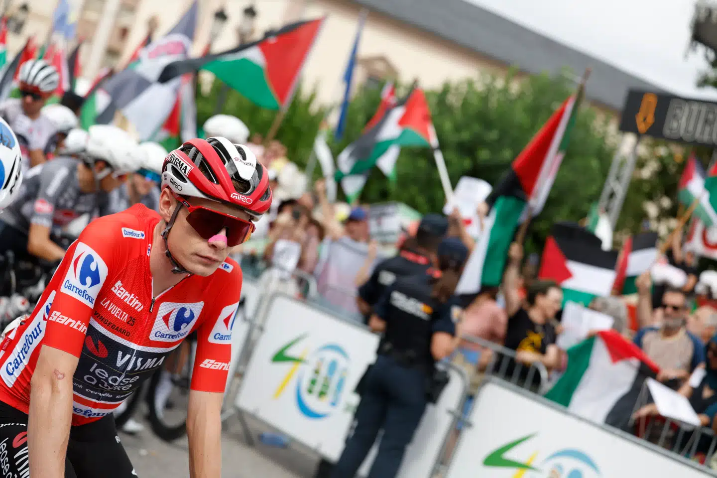 Jonas Vingegaard i løbets rødeførertrøje. I baggrunden ses tilskuere med det palæstinensiske flag.