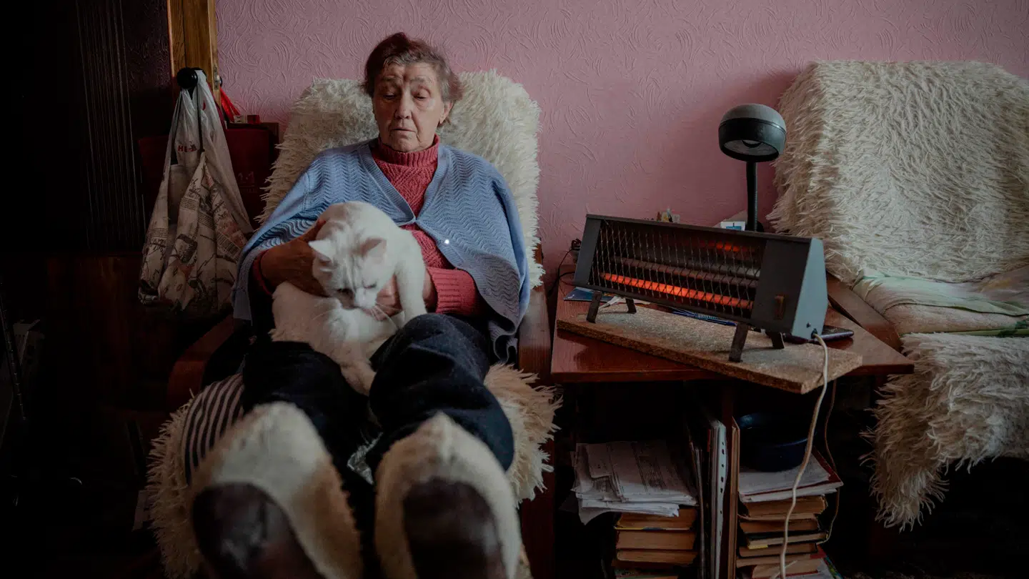 En ældre ukrainsk kvinde forsøger at holde varmen ved hjælp af store mængder tøj samt en varmelampe efter russiske angreb på Ukraines infrastruktur har lammet varmeproduktionen i store dele af landet.