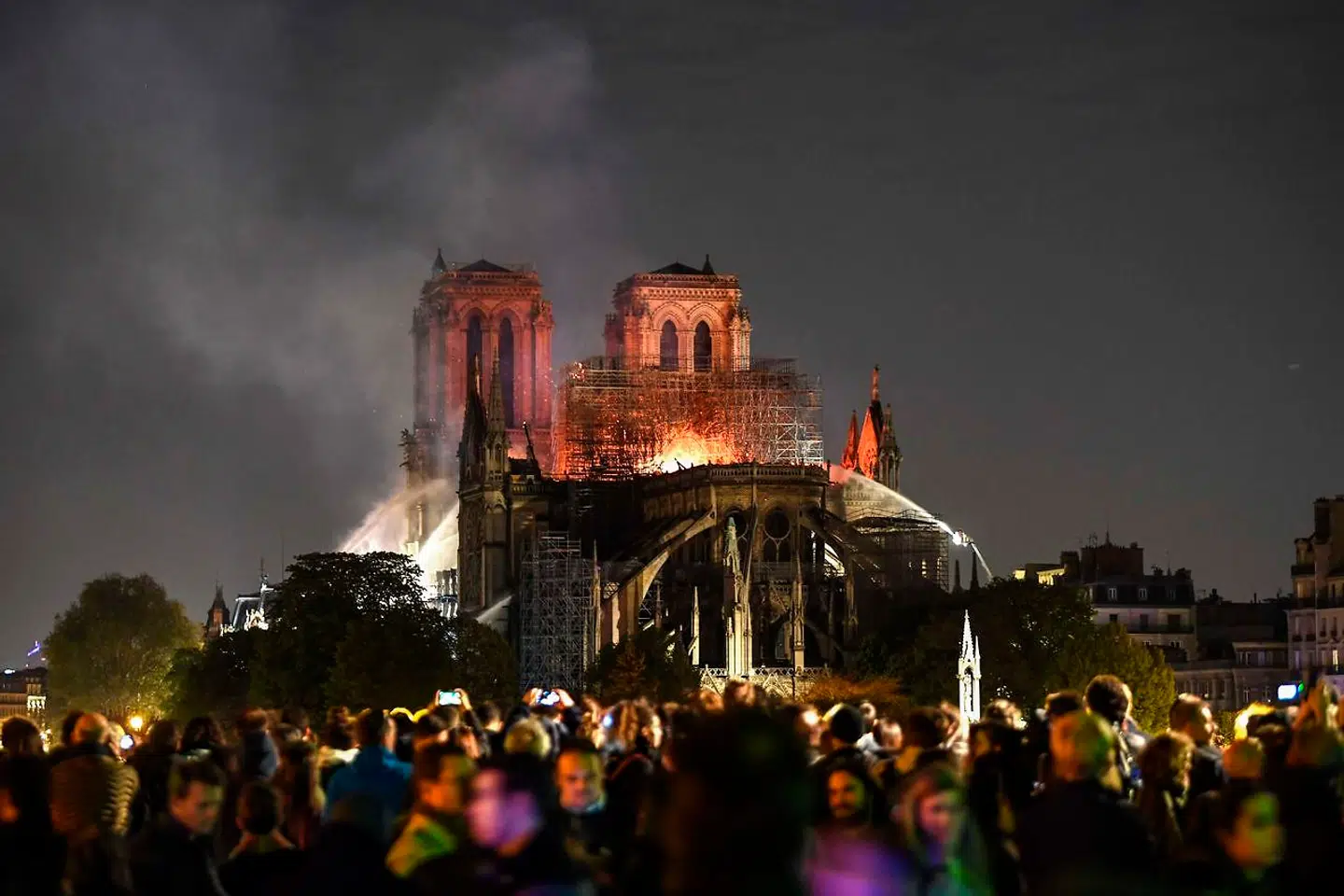 TOPSHOT - Bystanders look on as flames and smoke are seen billowing from the roof at Notre-Dame Cathedral in Paris on April 15, 2019. - A fire broke out at the landmark Notre-Dame Cathedral in central Paris, potentially involving renovation works being carried out at the site, the fire service said. Images posted on social media showed flames and huge clouds of smoke billowing above the roof of the gothic cathedral, the most visited historic monument in Europe. (Photo by ERIC FEFERBERG / AFP)