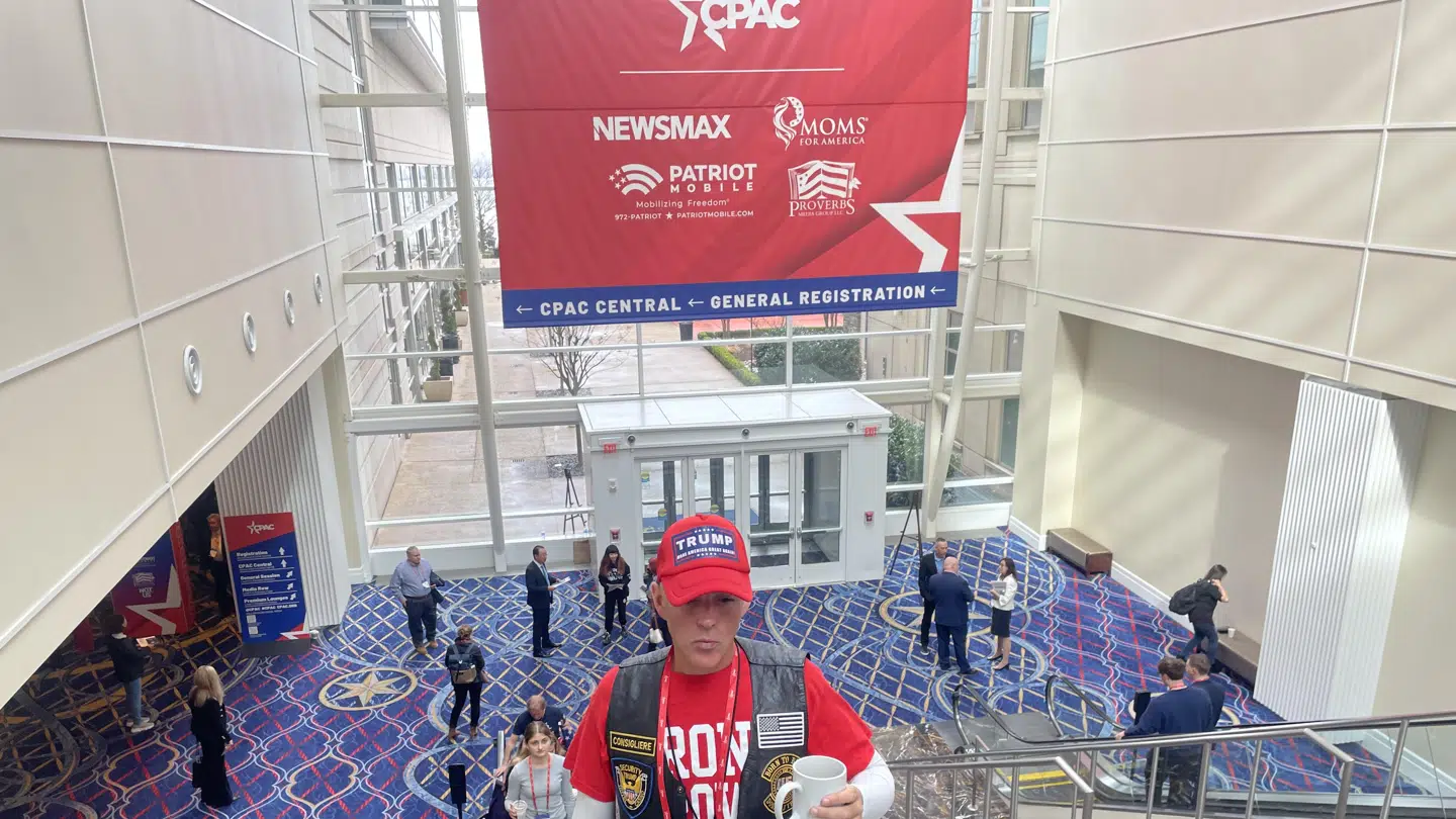 En Trump-tilhænger ved CPAC. Bag ham et enormt banner med CPACs partnere. Heriblandt Newsmax.