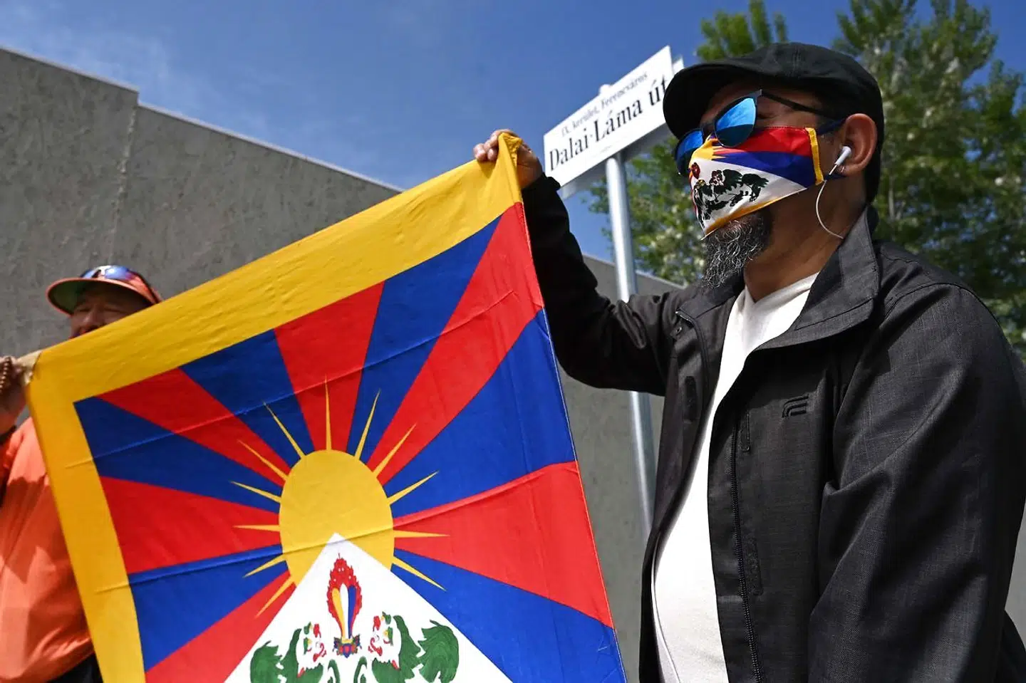 Aktivister holder det tibetanske flag oppe under et vejskilt med navnet 'Dalai Lama Street'.