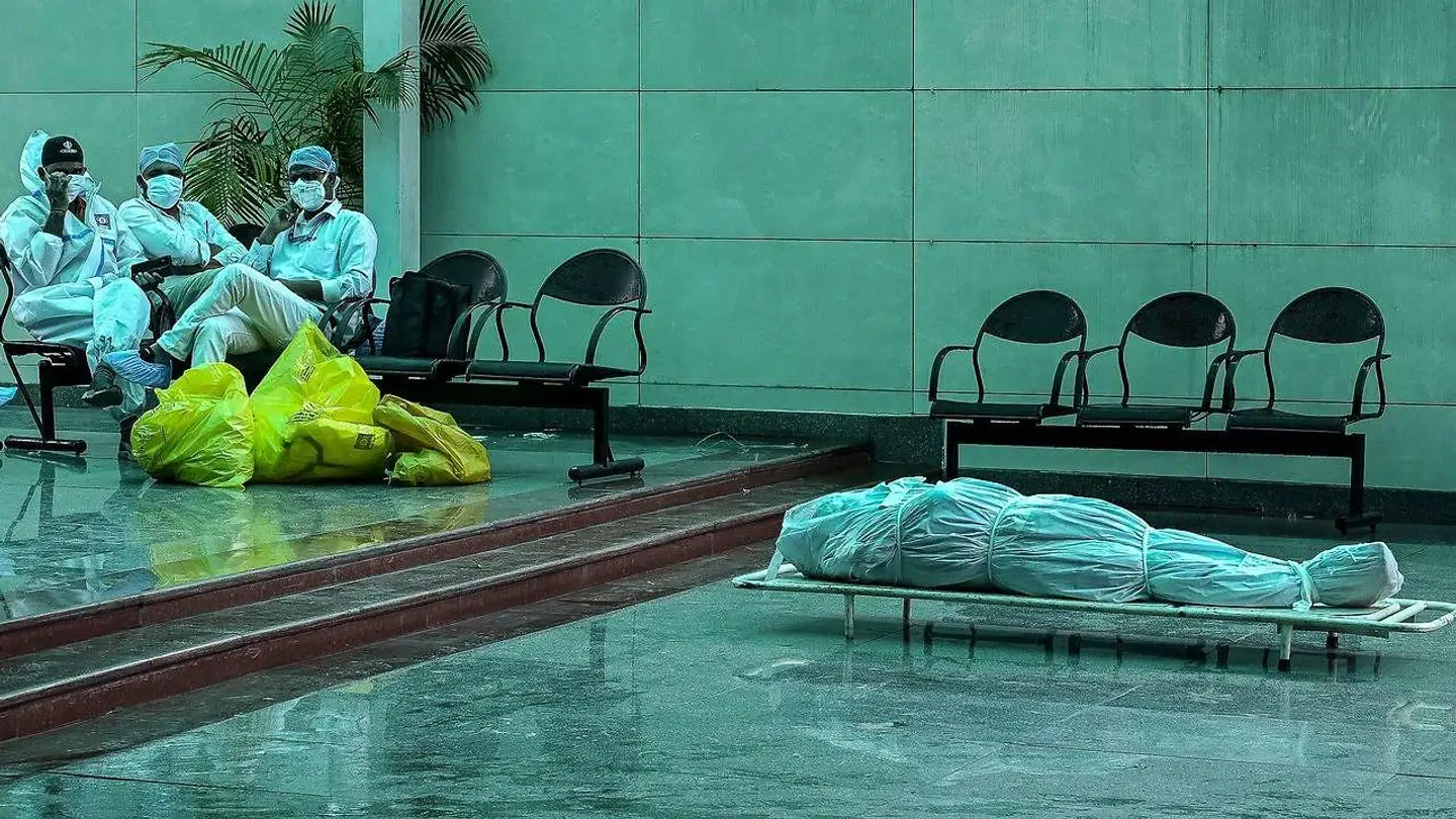Udmattet sundhedspersonale på et krematorium i New Delhi tager sig et hvil, mens en afdød med covid-19 ligger på en båre tæt ved.