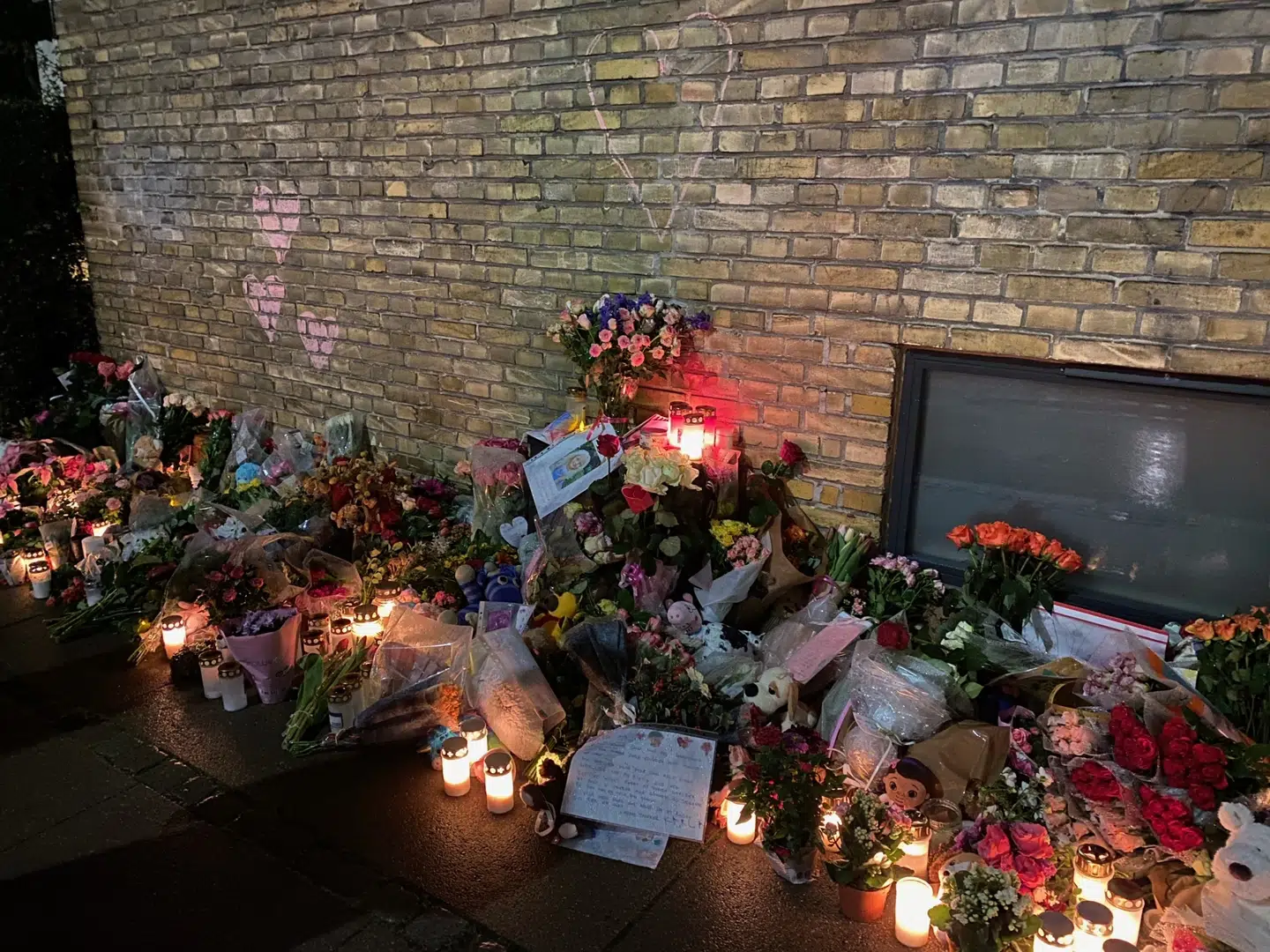 Der er tegnet røde kridthjerter på væggen. På fortovet står blomster, bamser og levende lys.