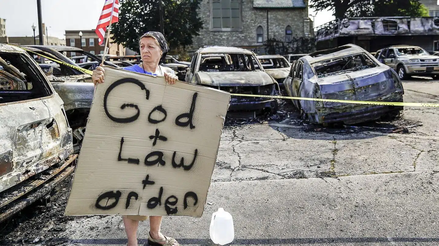En kvinde holder et amerikansk flag og et skilt, mens hun står ved siden af udbrændte biler efter en nat med voldsomme demonstrationer.