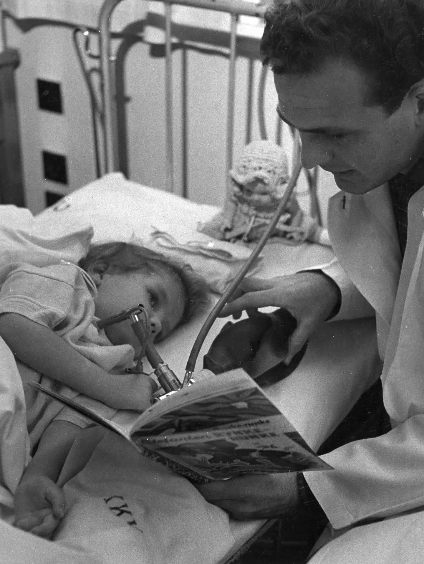 Overtryksventilation af et barn med lammelser i åndedrætsorganerne. Bemærk at den unge læge eller lægestuderende både udfører håndventilering og læser for pigen på samme tid. Polio blev også kaldt børnelammelse, fordi der var et forholdsmæssigt stort antal børn og unge der blev ramt. Foto: Medicinsk Museion, Københavns Universitet