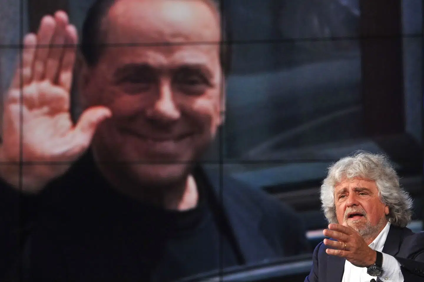 Femstjernebevægelsens leder, Beppe Grillo, tordner mod korruption under en tv-debat, mens Berlusconi smilende »ser til« fra en storskærm i studiet.