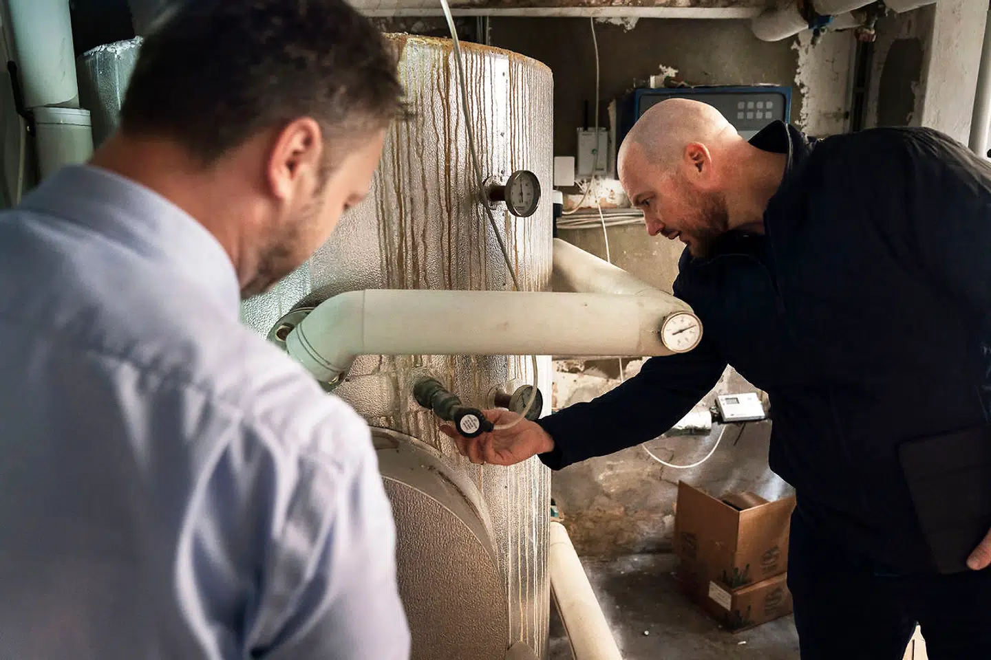 Bjørn Rhyding peger et klistermærke ud med et telefonnummer. »Nødhjælp, man ringer til, når det går galt,« forklarer han. Jeppe Blohm Nielsen supplerer: »Varmecentralen er ofte et glemt rum, indtil noget går galt.«