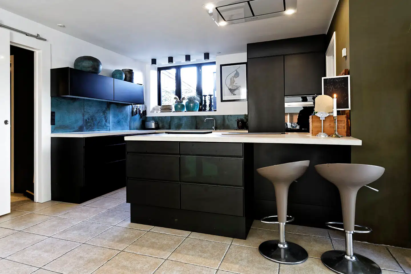 Det oprindeligt hvide Multiform-køkken fra 2005 er for nylig lakeret sort. Fliser på væggen er i petrol-turkis nuance. Søren Andersen går op i madlavning og har udskiftet den gamle pladskrævende emhætte med den nye loftsintegrerede fra Silverline.