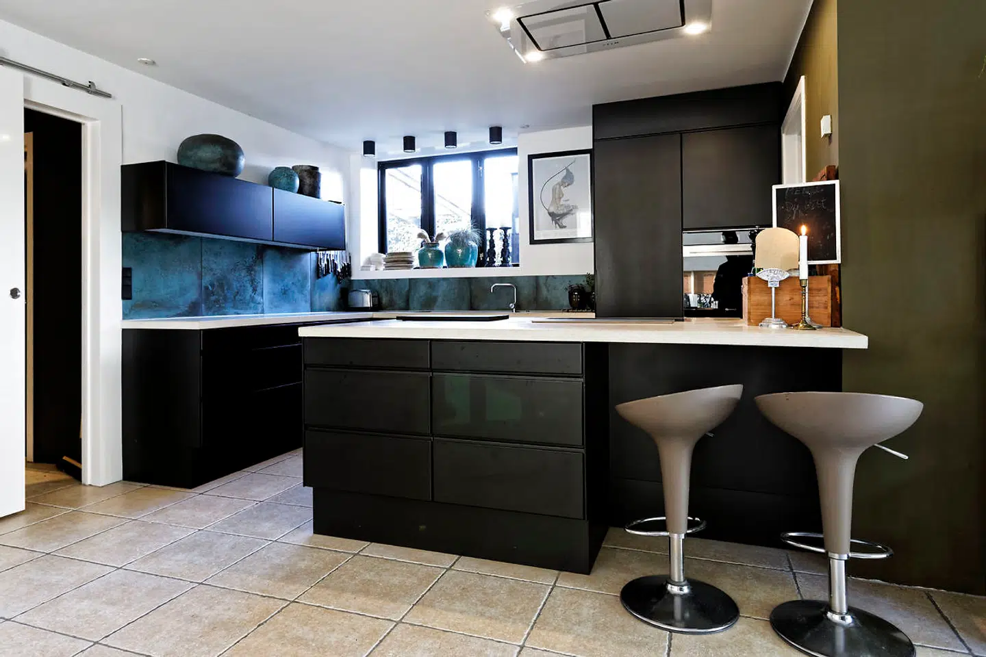 Det oprindeligt hvide Multiform-køkken fra 2005 er for nylig lakeret sort. Fliser på væggen er i petrol-turkis nuance. Søren Andersen går op i madlavning og har udskiftet den gamle pladskrævende emhætte med den nye loftsintegrerede fra Silverline.