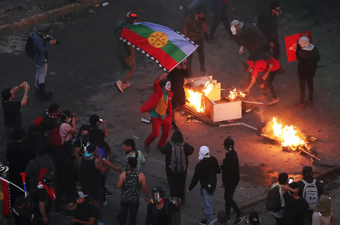 En demonstrant klædt som Joker bærer et flag ved siden af brændende genstande i Chiles hovedstad, Santiago, hvor demonstranter protesterer mod landets regering.