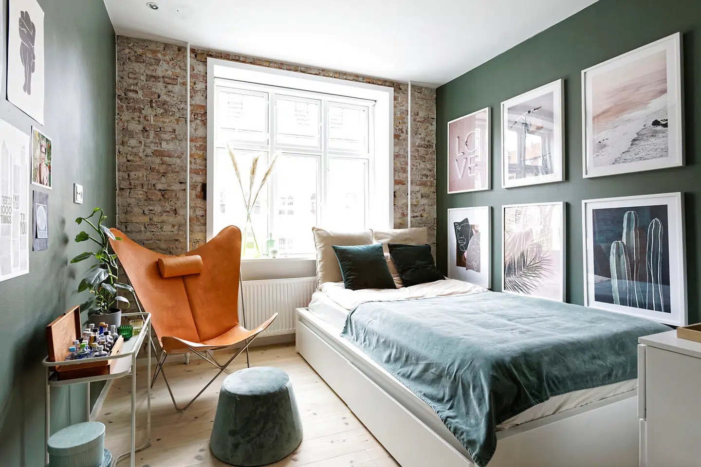 Soveværelset med den grå grønne vægfarve og den rå ydermur mod gaden var tidligere stue. Fremfor et klædeskab har Emilie Bjørnskov prioriteret plads til sin flagermusstol af kernelæder, en velourpuf og god gulvplads.