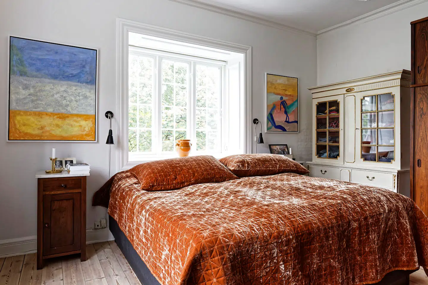 Soveværelset på første sal er fortsat med hvide vægge, selv om tapet har været overvejet. Det store vindue bag sengen har udsigt til en stor trækrone og er etableret for nylig. Det rustrøde veloursengetæppe er fra Ellos.