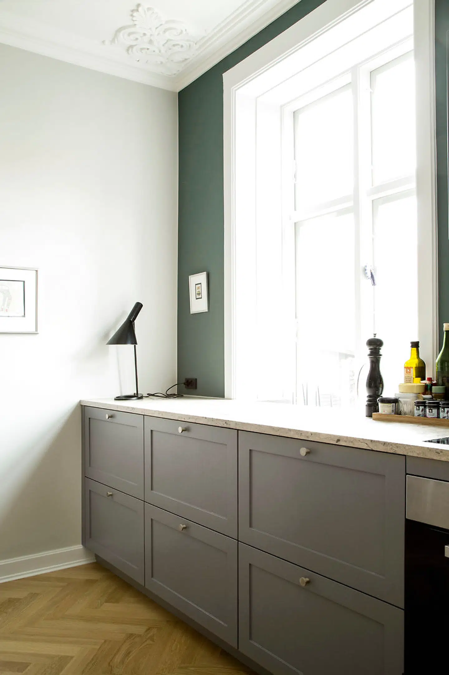 Køkkenet er skabt med tanke på harmoni, og derfor er skæringer og profiler på lågerne ens på det hele. Roen i rummet er vigtig, og derfor har parret valgt at male vægge og træværk lysegråt med farven Two Grey Rooms fra File Under Pop. Endevæggen med vinduer har dog fået en mørkere grøn, som giver kontrast og dybde til rummet. Den hedder Green Bananas og er også fra File Under Pop.