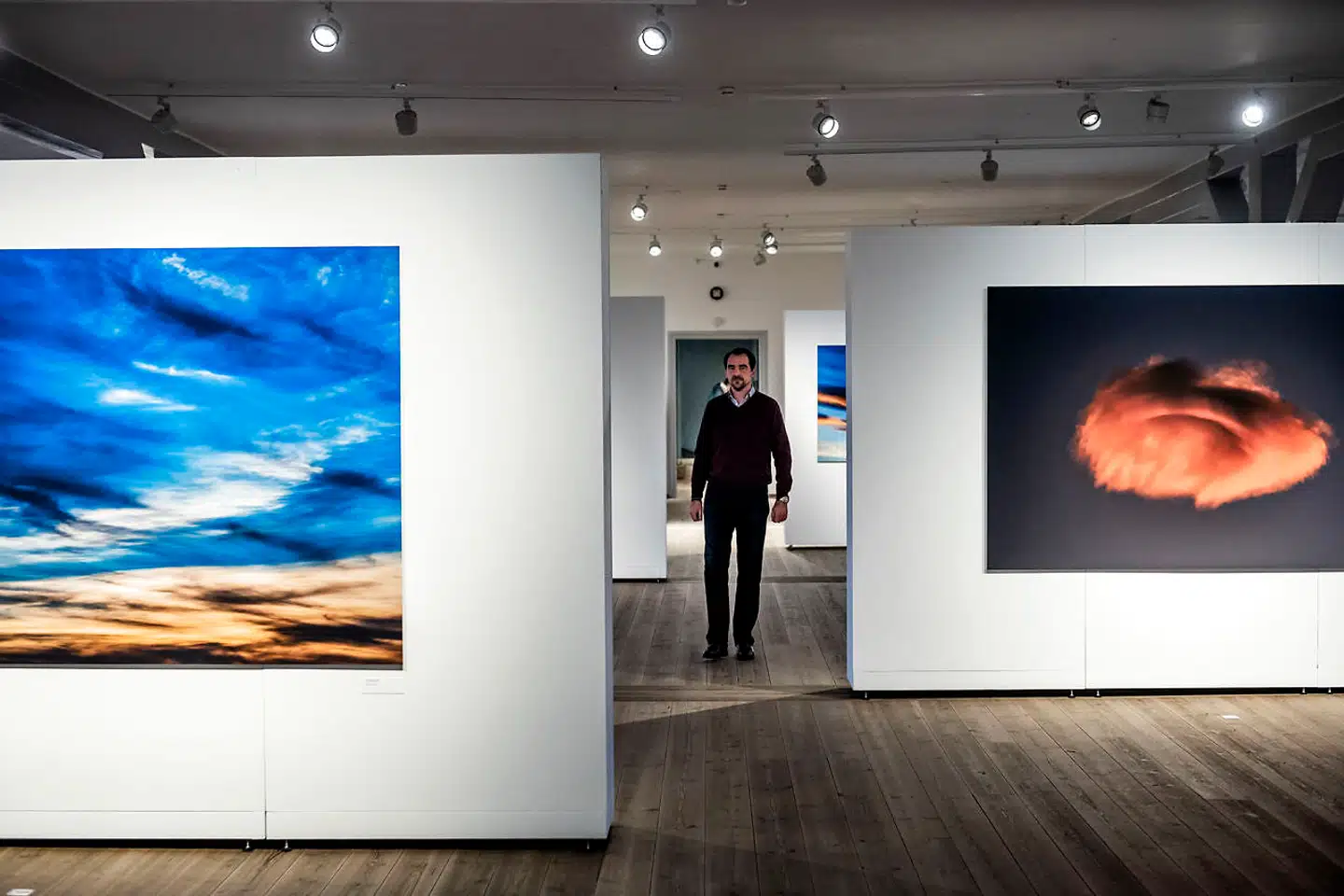 Prins Nikolaos på vej gennem udstillingen i Rundetaarn. Til venstre det billede, han har kaldt »Gratitude«. Billedet til højre har fået titlen »Hygge«. Prinsen havde ventet længe med kameraet parat for at fange et helt andet motiv, da den orange, pelsagtige sky pludselig dukkede op i hans synsfelt. »Det ligner sådan en ting, man kunne have lyst til at kramme eller krybe ind i, og egentlig havde jeg tænkte at kalde billedet »Cosy«. Men vi er jo i Danmark, smiler han.