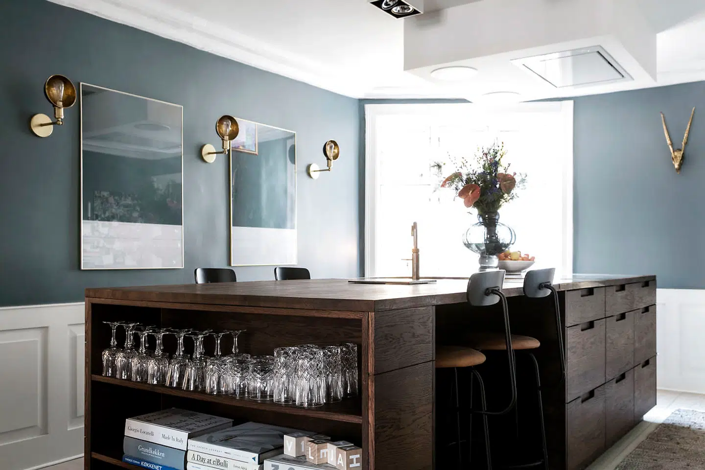 Køkkenet i røget eg er tegnet af Camilla Wikke Wahlstrøm selv og bygget af Handcrafted Interior. Messingvasken, som var hele udgangspunktet for køkkenet, er også specialbygget og fra Ono. Billederne på væggen er billeder af væggen, som er blevet indrammet. Barstolene er fra Menu, ligesom væglamperne, som er blevet vendt både opad og udad for at give dem et andet udtryk.