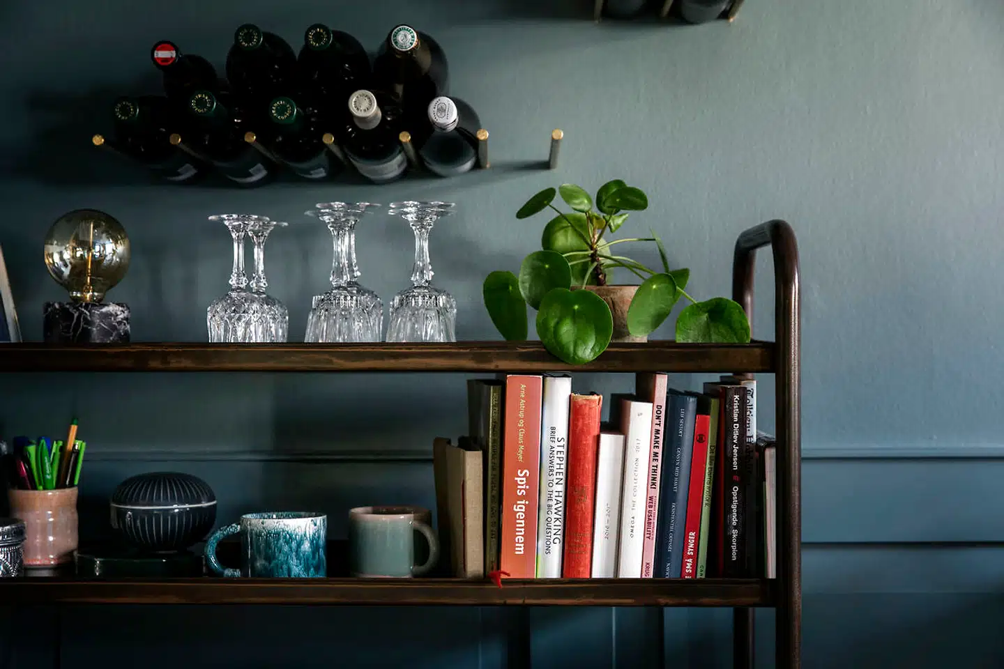Reolen i i bruneret messing er fra Rue Verte, og her står kopper fra Studio Arhoj, smukke glas og kogebøger. Vinstængerne fra Erbs Studio har parret fået sat ind i væggen, fordi deres lille datter blev ved med at hive flaskerne ud af reolen.