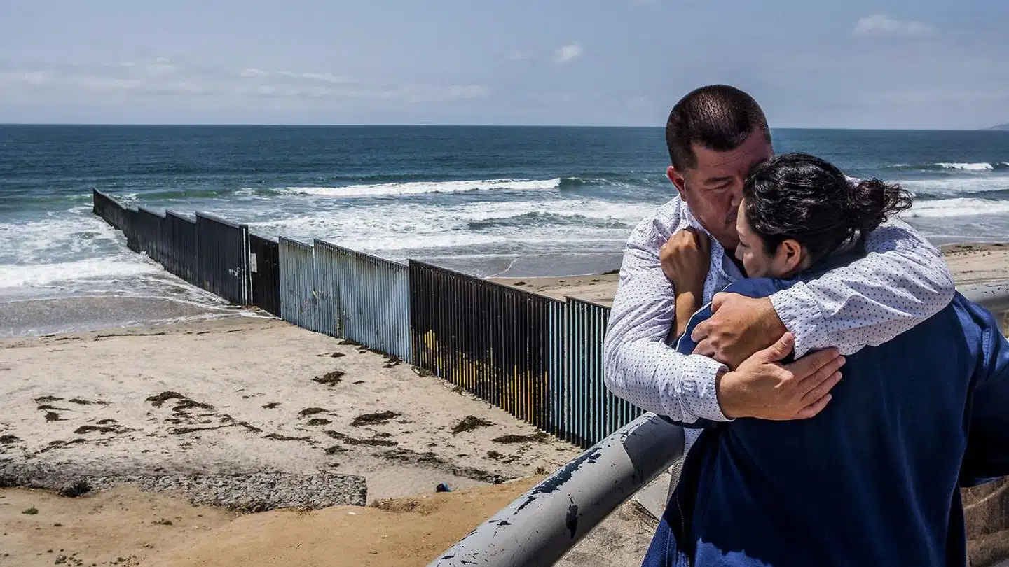 En mand krammer og trøster sin kone ved det høje hegn på grænsen, der fortsætter ud i stillehavet, mellem USA og Mexico. I dag er der et op til seks meter højt hegn langs 1.100 km af den 3.200 km lange grænse, omfattende elektronisk overvågning og tusindvis af patruljerende grænsebetjente. Trump forlanger at få en mur på hele grænsen.