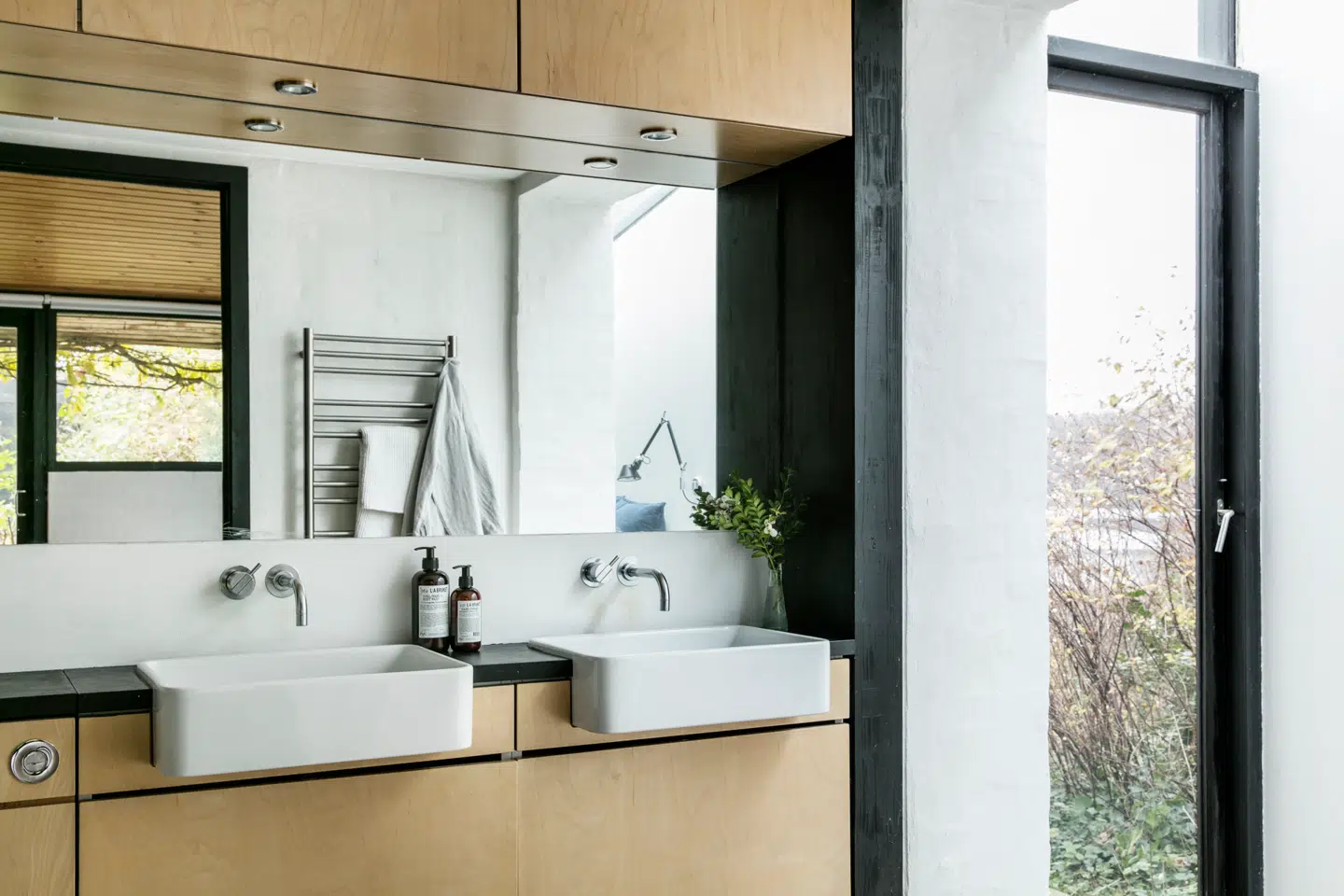 Det er Nikolaj Ernst selv, der har tegnet badeværelset, og snedkeren Søren Risvang har bygget træskabene i badeværelset. I spejlet kan man ane sengelampen og vinduerne i soveværelset, så rummene hænger sammen, men er stadig adskilt.