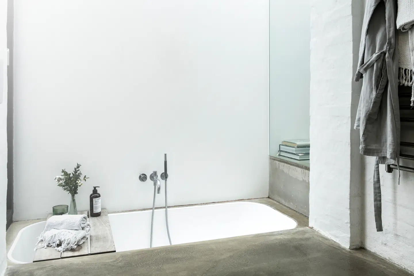 Parret fik gravet ud, så de kunne sænke badekarret, så det flugter med gulvet og samtidig gør, at man har fri udsigt til søen fra både sengen og badet. Camilla Gullits bryder det hårde udtryk med tekstiler og organisk tilbehør. Bemærk overgangen mellem badeværelset og soveværelset til højre i billedet.