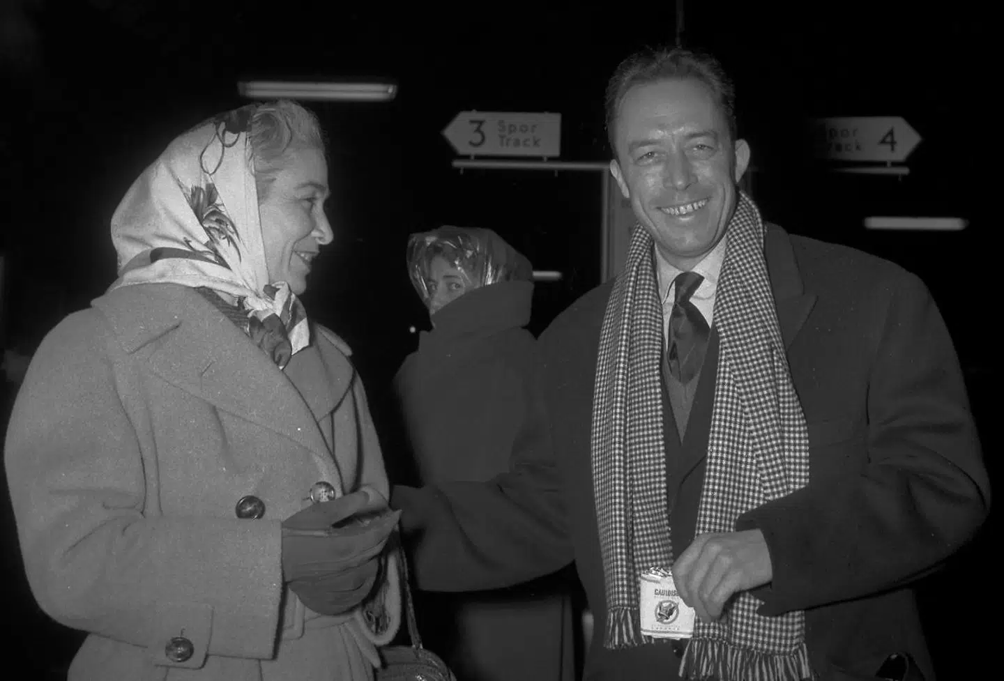 »Aarets litterære Nobel-pristager, den 44-aarige franske forfatter Albert Camus, kom i aftes til København med Nordeekspressen og tilbragte en times tid her, før han tog videre nordpaa til Stockholm, hvor han skal have overrakt den fornemme pris,« skriver Berlingske Tidende mandag den 9. december 1957. Albert Camus var bl.a. ledsaget af sin ægtefælle Francine Camus (tv). I Camus' højre hånd hans trofaste Gauloises. Foto: Hakon Nielsen