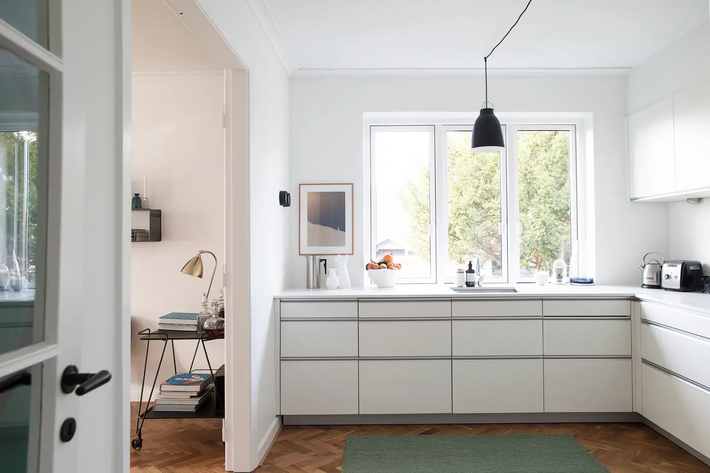 Køkkenet er indrettet med lyse vægge og lofter for at udnytte dagslyset, der kommer ind ad det store vinduesparti. Parret har piftet den enkle og stilrene køkkenløsning fra Alno op med kunst på væggen, mens gulvet har fået selskab af et stort, grønt gulvtæppe fra Muuto. Derover svæver en pendel fra Lightyears.