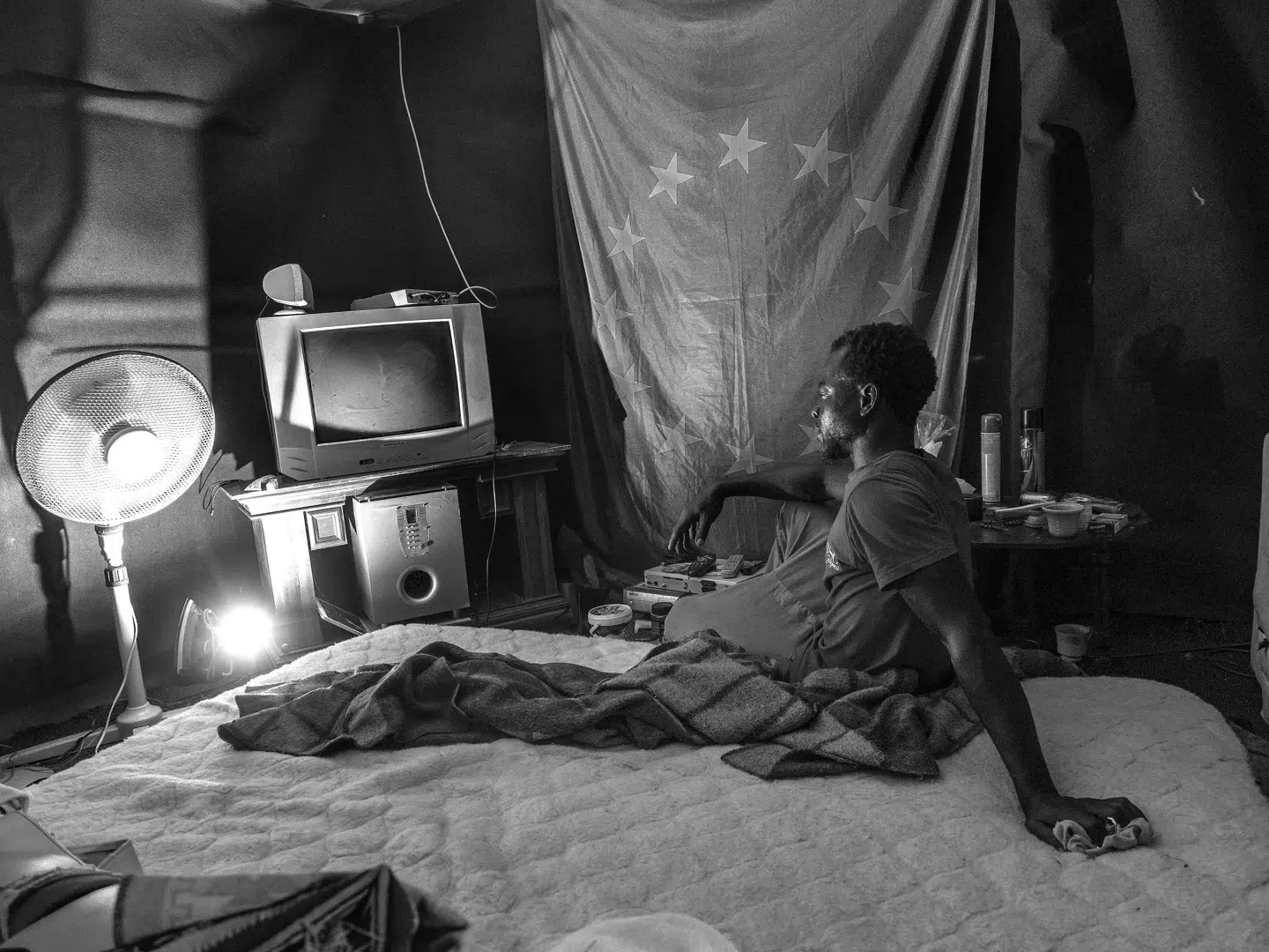 36-årige Sofo fra Ghana. Var fisker hjemme i Ghana, har nu boet i migrantlejren i to år og lever af »forskellige jobs«. Om EU-flaget på væggen siger han: »Ved ikke, hvad det er for et flag, men jeg kan lide det, og derfor hængte jeg det op.« FOTO: ENRI CANAJ, MAGNUM